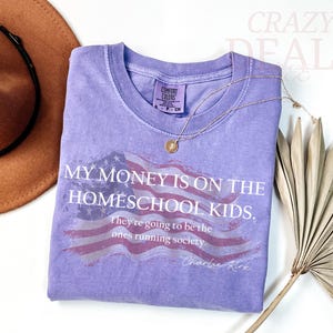 Pu&ograve; includere: Una maglietta lilla Comfort Colors con la scritta "MY MONEY IS ON THE HOMESCHOOL KIDS." e un disegno della bandiera americana. Una collana d'oro e un cappello marrone sono anche nell'immagine.