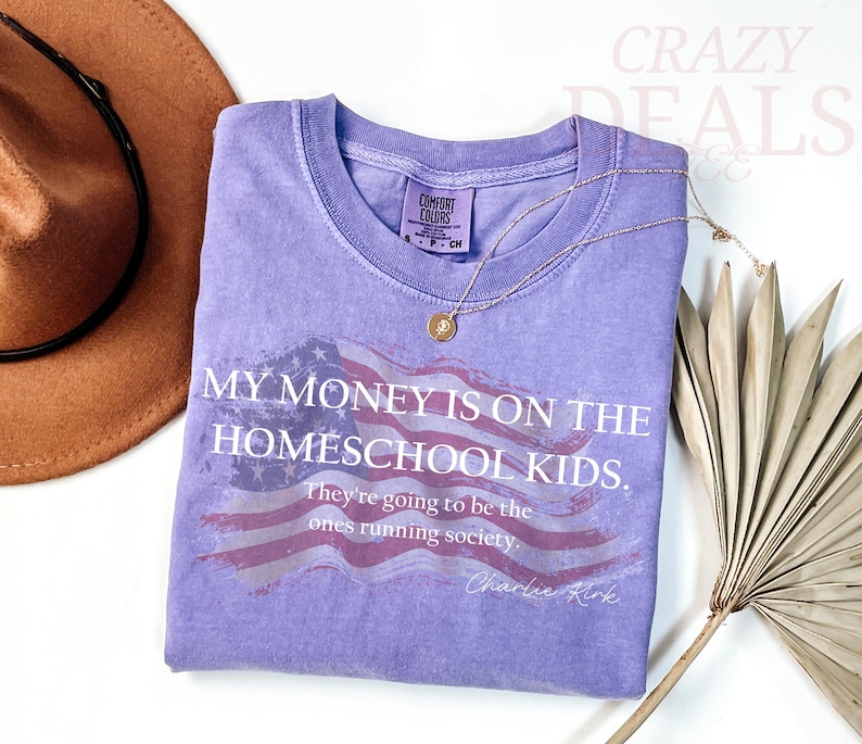 Pu&ograve; includere: Una maglietta lilla Comfort Colors con la scritta "MY MONEY IS ON THE HOMESCHOOL KIDS." e un disegno della bandiera americana. Una collana d'oro e un cappello marrone sono anche nell'immagine.