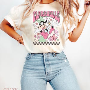 May include: Cream-coloured t-shirt with a retro graphic of Clarabelle Cow in a pink outfit, surrounded by flowers and the text "CLARABELLE" in pink. The design includes a checkered border at the bottom. The shirt is paired with blue jeans.