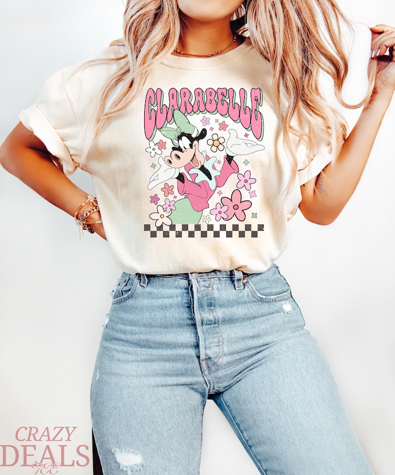 May include: Cream-coloured t-shirt with a retro graphic of Clarabelle Cow in a pink outfit, surrounded by flowers and the text "CLARABELLE" in pink. The design includes a checkered border at the bottom. The shirt is paired with blue jeans.