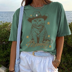 May include: Green t-shirt featuring a cat wearing a cowboy hat and bandana, with the word "Meowdy" in a script font. The design includes gold stars. The shirt is paired with white shorts and a blue belt.