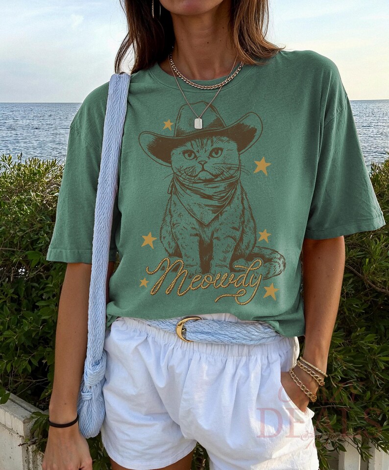 May include: Green t-shirt featuring a cat wearing a cowboy hat and bandana, with the word "Meowdy" in a script font. The design includes gold stars. The shirt is paired with white shorts and a blue belt.