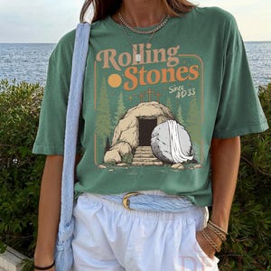 Pu&ograve; includere: T-shirt verde con la scritta "Rolling Stones Since 4D 33" in arancione e bianco. La grafica raffigura una tomba con croci, un sole e una pietra. La maglietta &egrave; abbinata a pantaloncini bianchi e una borsa blu.