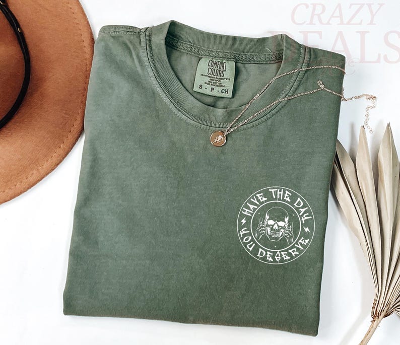 Puede incluir: Camiseta verde oliva con cuello redondo y mangas cortas. La parte trasera de la camiseta tiene un gr&aacute;fico blanco con una calavera y el texto "HAVE THE DAY YOU DESERVE". Un collar dorado con un colgante est&aacute; sobre la camiseta.