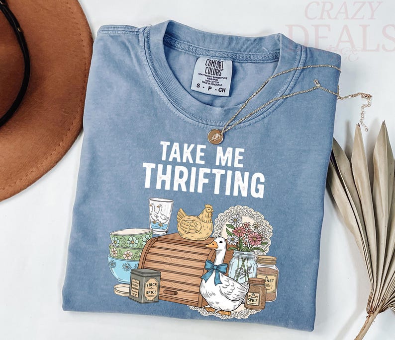 以下が含まれることがあります： 「TAKE ME THRIFTING」の文字とアヒル、鶏、パン箱、瓶のグラフィックが描かれた、ライトブルーのComfort Colors Tシャツ。 金色のネックレスがシャツの上にかけられています。