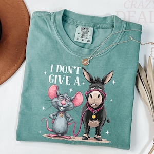 Puede incluir: Una camiseta Comfort Colors verde azulado con un ratón y un burro de dibujos animados y el texto "I DON'T GIVE A". El ratón es gris con orejas rosas y una estrella. El burro es negro con un cabestro rosa.