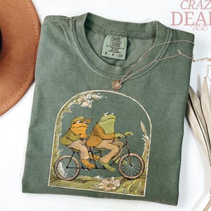 May include: Olive green Comfort Colors t-shirt featuring a whimsical illustration of two frogs riding a tandem bicycle. The graphic is framed by an arch with floral accents. A gold necklace and a brown hat are also visible.