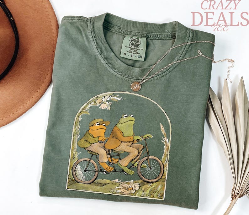 May include: Olive green Comfort Colors t-shirt featuring a whimsical illustration of two frogs riding a tandem bicycle. The graphic is framed by an arch with floral accents. A gold necklace and a brown hat are also visible.