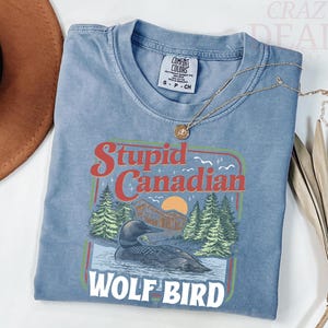 Puede incluir: Una camiseta azul Comfort Colors con un gr&aacute;fico que dice "Stupid Canadian Wolf Bird". El gr&aacute;fico presenta un somorgujo en el agua, &aacute;rboles y una puesta de sol. Un collar de oro est&aacute; drapeado sobre la camisa.