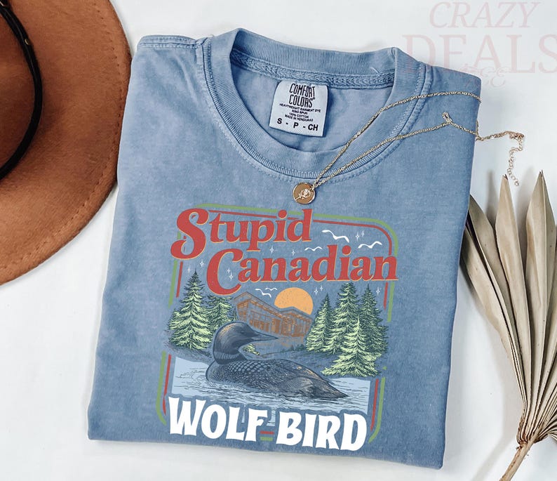 Puede incluir: Una camiseta azul Comfort Colors con un gr&aacute;fico que dice "Stupid Canadian Wolf Bird". El gr&aacute;fico presenta un somorgujo en el agua, &aacute;rboles y una puesta de sol. Un collar de oro est&aacute; drapeado sobre la camisa.