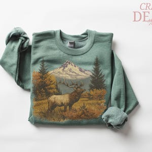 May include: A sage green sweatshirt with a detailed graphic of an elk in a mountain landscape. The design includes a mountain range, trees, and autumn foliage. The text "CRAZY DEALS" is in the upper right corner.