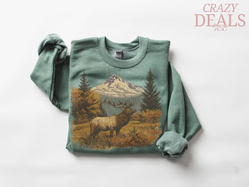 May include: A sage green sweatshirt with a detailed graphic of an elk in a mountain landscape. The design includes a mountain range, trees, and autumn foliage. The text "CRAZY DEALS" is in the upper right corner.