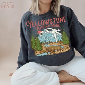 May include: Dark gray sweatshirt featuring a graphic of Yellowstone National Park. The design includes mountains, trees, a river, and a bison, with the text "YELLOWSTONE NATIONAL PARK" in a vintage style.