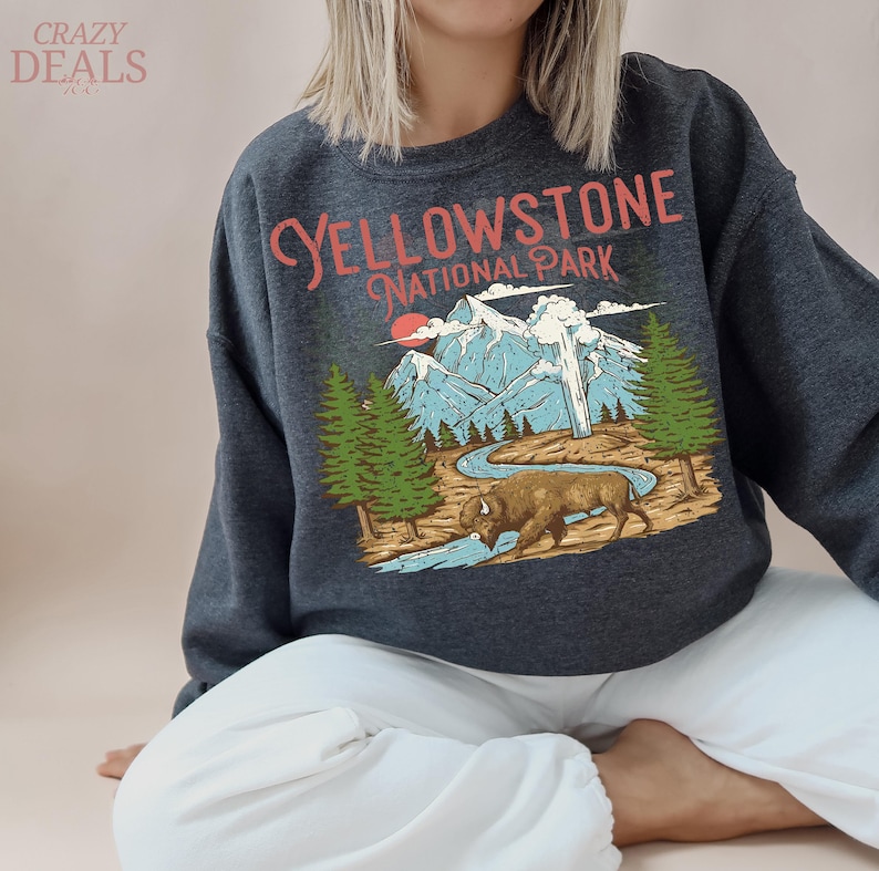 May include: Dark gray sweatshirt featuring a graphic of Yellowstone National Park. The design includes mountains, trees, a river, and a bison, with the text "YELLOWSTONE NATIONAL PARK" in a vintage style.