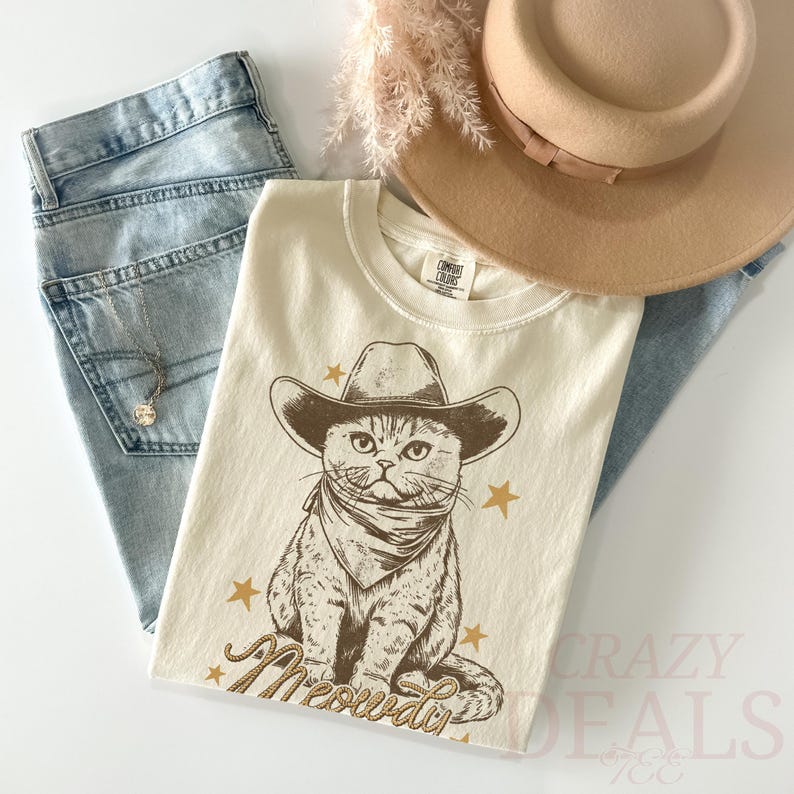 May include: Cream-colored t-shirt featuring a cartoon cat wearing a cowboy hat and bandana, with the word "Meowdy" below. The shirt is paired with light-wash jeans, a tan hat, and a necklace.