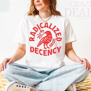 May include: White t-shirt with a red graphic that reads "RADICALIZED BY BASIC DECENCY" around a bird holding a branch. The person is wearing light blue jeans and white sneakers.