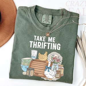 以下が含まれることがあります： 「TAKE ME THRIFTING」の白い文字が入ったオリーブグリーンのTシャツ。青いリボンをつけたアヒル、鶏、パン箱、スパイスジャーのグラフィック。金のネックレスがシャツの上にかけられています。