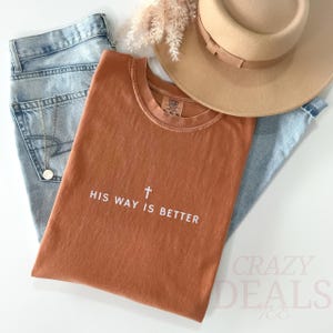 May include: A terracotta-coloured t-shirt with the text "HIS WAY IS BETTER" and a cross graphic. The shirt is styled with light blue jeans, a beige hat, and a gold necklace. The image has a casual, lifestyle aesthetic.