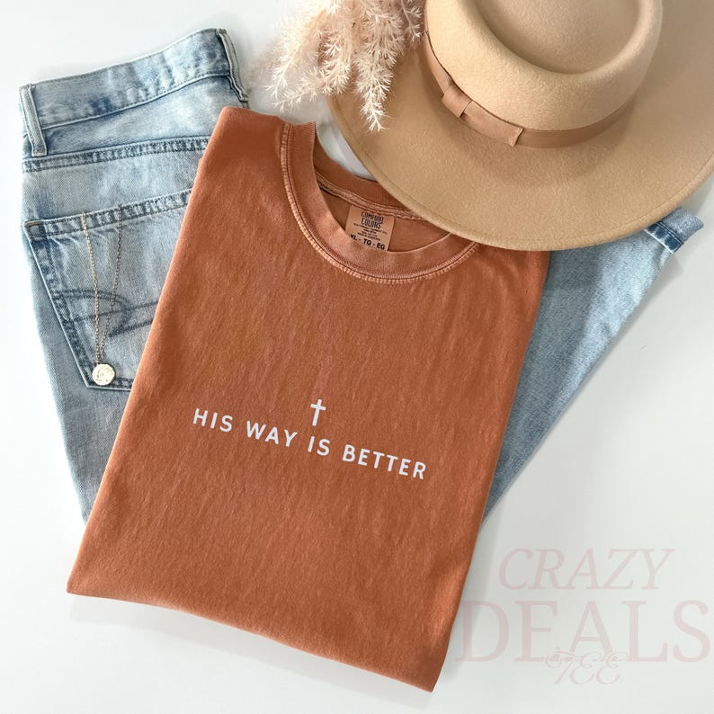 May include: A terracotta-coloured t-shirt with the text "HIS WAY IS BETTER" and a cross graphic. The shirt is styled with light blue jeans, a beige hat, and a gold necklace. The image has a casual, lifestyle aesthetic.