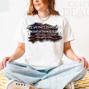 Pu&ograve; includere: T-shirt bianco con la scritta "MY MONEY IS ON THE HOMESCHOOL KIDS" e "They're going to be the ones running society" in nero. Il design include una bandiera americana invecchiata.