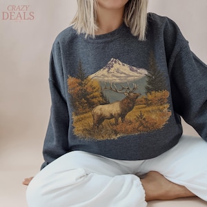 May include: Dark grey sweatshirt with a mountain and elk graphic. The design includes a mountain, trees, and an elk in a field. The text "CRAZY DEALS" is in the upper left corner. The sweatshirt is a casual, comfortable style.