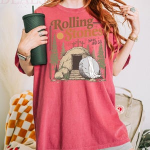 Pu&ograve; includere: T-shirt rossa con una grafica con le parole "Rolling Stones" e un'illustrazione di una tomba con una grande pietra. La maglietta &egrave; di colore rosso pieno e ha maniche corte. Un bicchiere verde scuro &egrave; tenuto nella mano sinistra.
