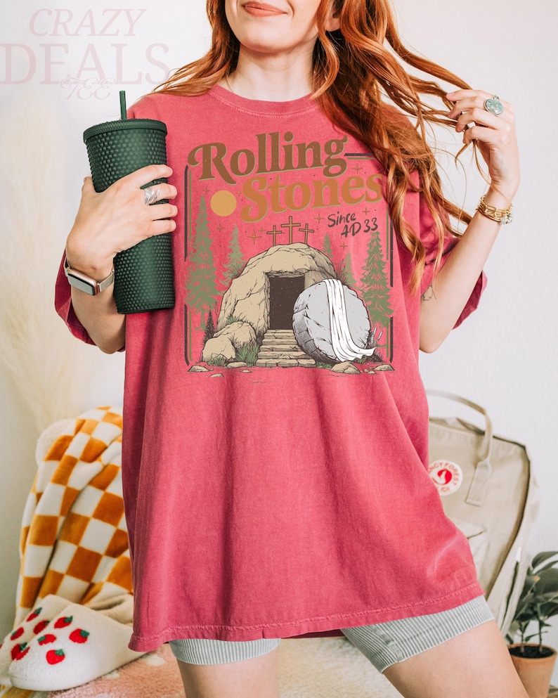 Pu&ograve; includere: T-shirt rossa con una grafica con le parole "Rolling Stones" e un'illustrazione di una tomba con una grande pietra. La maglietta &egrave; di colore rosso pieno e ha maniche corte. Un bicchiere verde scuro &egrave; tenuto nella mano sinistra.