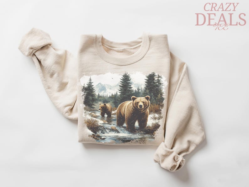 May include: Beige sweatshirt featuring a forest scene with two brown bears in a stream, surrounded by trees and mountains. The sweatshirt has long sleeves and a crew neck. The words "CRAZY DEALS" are in the upper right corner.