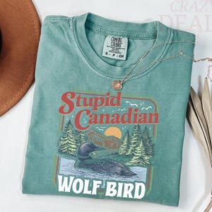 Puede incluir: Una camiseta Comfort Colors verde azulado con las palabras "Stupid Canadian Wolf Bird" en un dise&ntilde;o retro. El gr&aacute;fico incluye un somorgujo, &aacute;rboles y una puesta de sol. Un collar de oro descansa sobre la camisa.