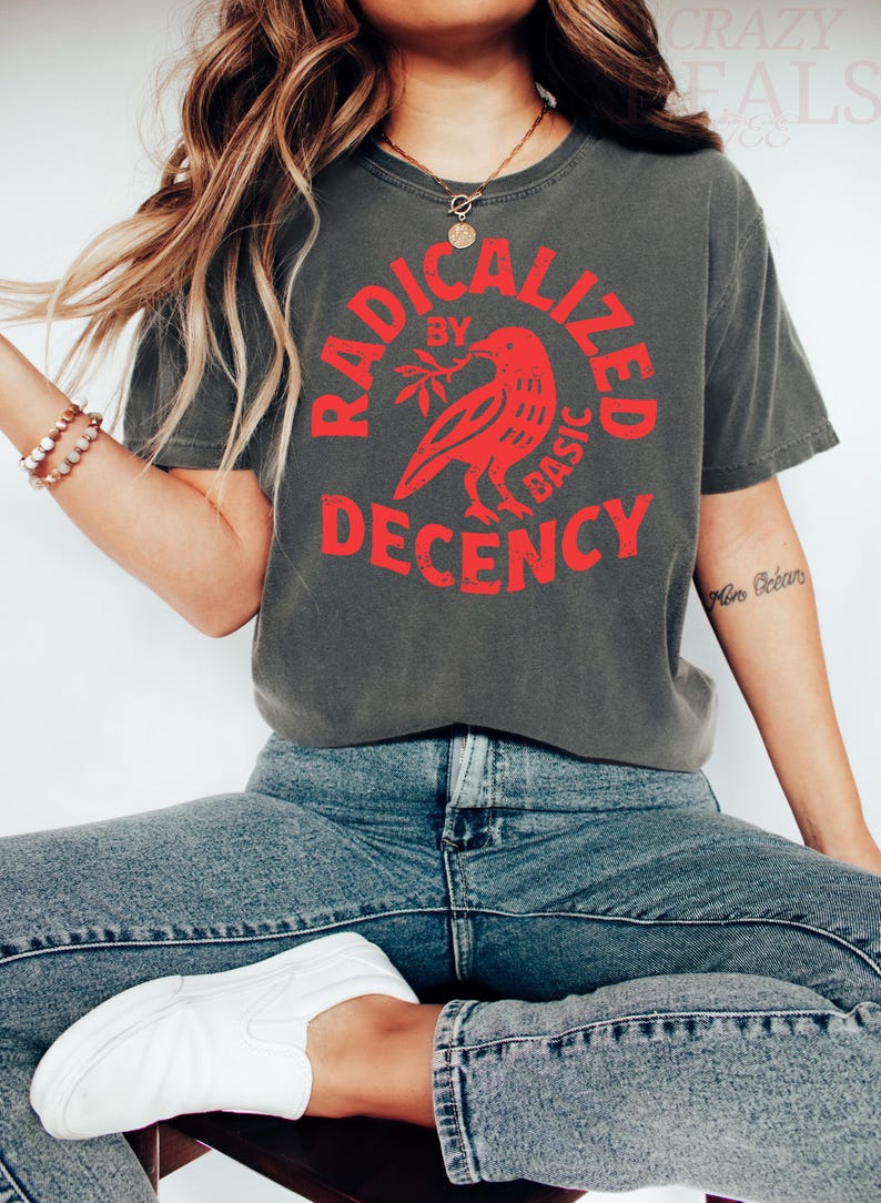 May include: Dark gray t-shirt with the red text "RADICALIZED BY BASIC DECENCY" and a red bird graphic. The shirt is paired with blue jeans and white sneakers. A necklace and bracelets are also visible.