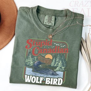 Puede incluir: Camiseta verde oliva con un gr&aacute;fico que presenta un somorgujo en un lago, pinos y el texto "Stupid Canadian Wolf Bird". Un collar dorado descansa sobre la camisa. La camisa est&aacute; doblada y se muestra con un sombrero marr&oacute;n y una hoja de palma seca.