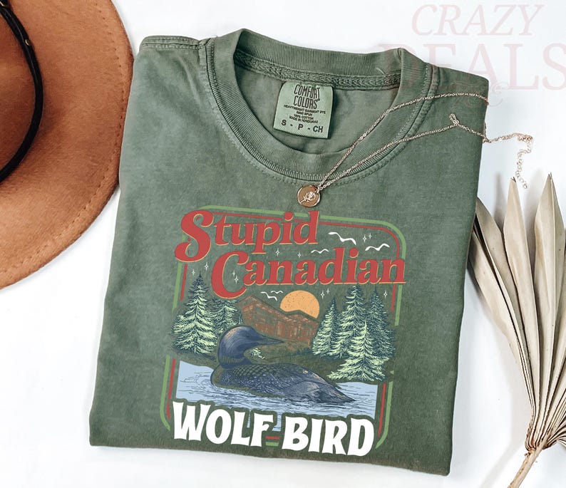 Puede incluir: Camiseta verde oliva con un gr&aacute;fico que presenta un somorgujo en un lago, pinos y el texto "Stupid Canadian Wolf Bird". Un collar dorado descansa sobre la camisa. La camisa est&aacute; doblada y se muestra con un sombrero marr&oacute;n y una hoja de palma seca.