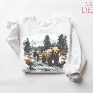 May include: White long-sleeved sweatshirt featuring a watercolor illustration of two brown bears in a forest stream. The design includes trees, mountains, and the text "CRAZY DEALS".