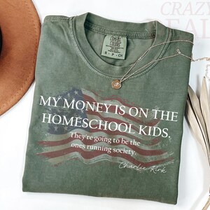 Pu&ograve; includere: Una maglietta verde oliva con la scritta "MY MONEY IS ON THE HOMESCHOOL KIDS" e un disegno della bandiera americana. Una collana d'oro con un ciondolo poggia sulla maglietta. Un cappello marrone &egrave; visibile nell'angolo in alto a sinistra.
