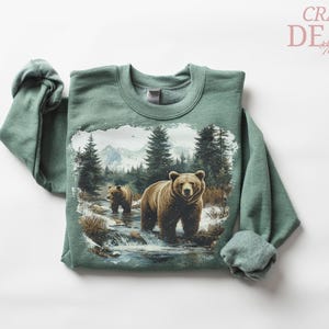 May include: A green sweatshirt featuring a detailed graphic of two brown bears in a snowy forest scene. The bears are standing in a stream with trees and mountains in the background. The text "CRAZY DEALS" is in the upper right corner.