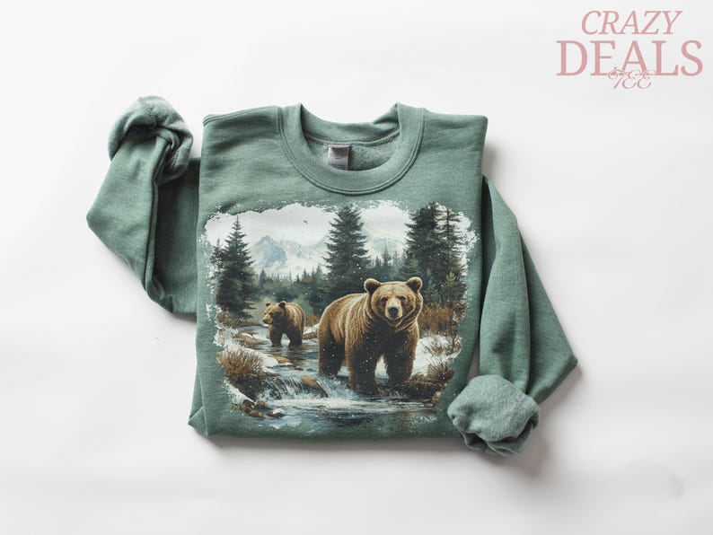 May include: A green sweatshirt featuring a detailed graphic of two brown bears in a snowy forest scene. The bears are standing in a stream with trees and mountains in the background. The text "CRAZY DEALS" is in the upper right corner.