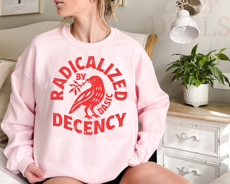 May include: A light pink sweatshirt with the words "RADICALIZED BY BASIC DECENCY" in red, encircling a red bird holding a sprig. The sweatshirt is a long-sleeved crew neck.