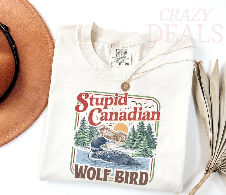 May include: Cream-colored t-shirt with a graphic that reads "Stupid Canadian Wolf Bird." The design features a loon on water, trees, a building, and a sunset. The shirt has a crew neck and a small tag.