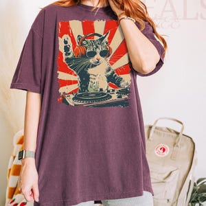 May include: A maroon oversized t-shirt with a graphic of a cat DJ wearing sunglasses and headphones, set against a red and cream sunburst design. The cat is behind a turntable. The text "Crazy Deals" is visible in the background.
