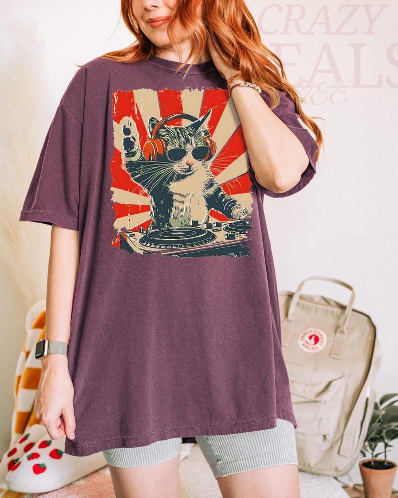 May include: A maroon oversized t-shirt with a graphic of a cat DJ wearing sunglasses and headphones, set against a red and cream sunburst design. The cat is behind a turntable. The text "Crazy Deals" is visible in the background.