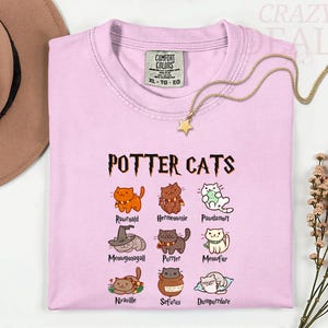 Pu&ograve; includere: T-shirt rosa chiaro con la scritta "POTTER CATS" e illustrazioni di gatti dei cartoni animati. La maglietta presenta vari personaggi di gatti con nomi come "Rawrnald" e "Hermeownie". Una collana d'oro con un ciondolo a forma di stella &egrave; drappeggiata sulla maglietta.