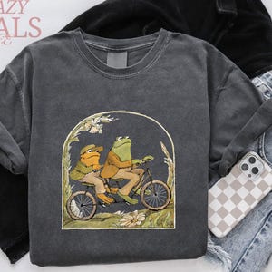 May include: Dark gray sweatshirt featuring a whimsical illustration of two frogs riding a bicycle. The artwork is framed by an arch with floral accents. The words "CRAZY DEALS" are printed at the top left.