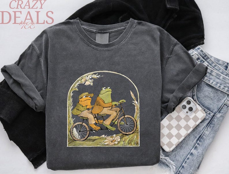 May include: Dark gray sweatshirt featuring a whimsical illustration of two frogs riding a bicycle. The artwork is framed by an arch with floral accents. The words "CRAZY DEALS" are printed at the top left.