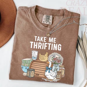 以下が含まれることがあります： 「TAKE ME THRIFTING」の文字とアヒル、鶏、花、その他のヴィンテージアイテムのグラフィックが描かれた茶色のTシャツ。シルバーのネックレスがシャツの上にかけられています。シャツのサイズはSです。