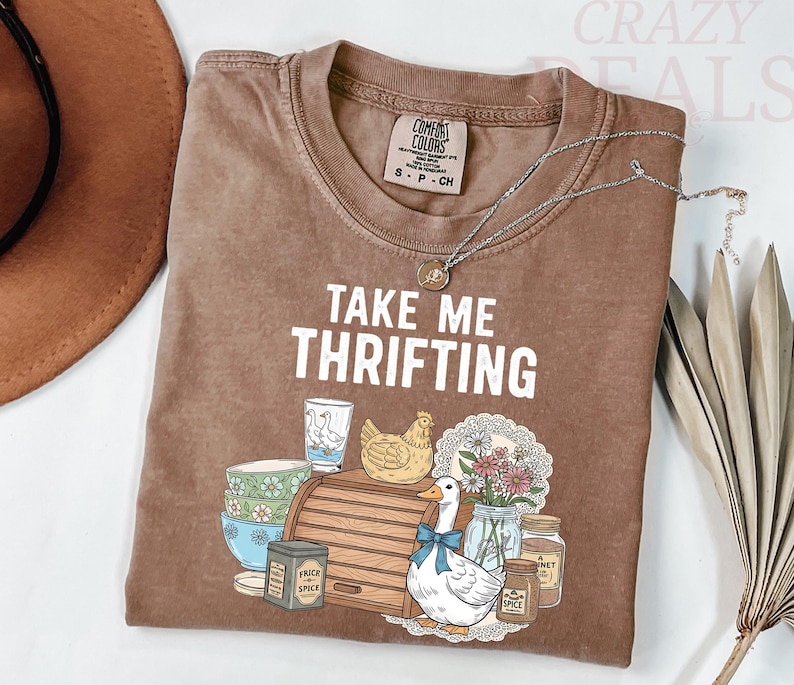 以下が含まれることがあります： 「TAKE ME THRIFTING」の文字とアヒル、鶏、花、その他のヴィンテージアイテムのグラフィックが描かれた茶色のTシャツ。シルバーのネックレスがシャツの上にかけられています。シャツのサイズはSです。