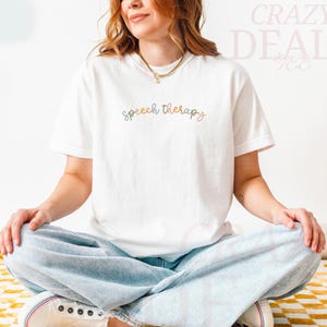 May include: White t-shirt with the words "speech therapy" in a cursive, multi-colored font. The shirt is worn with light blue jeans and white sneakers. The person is sitting cross-legged on a yellow and white checkered surface.