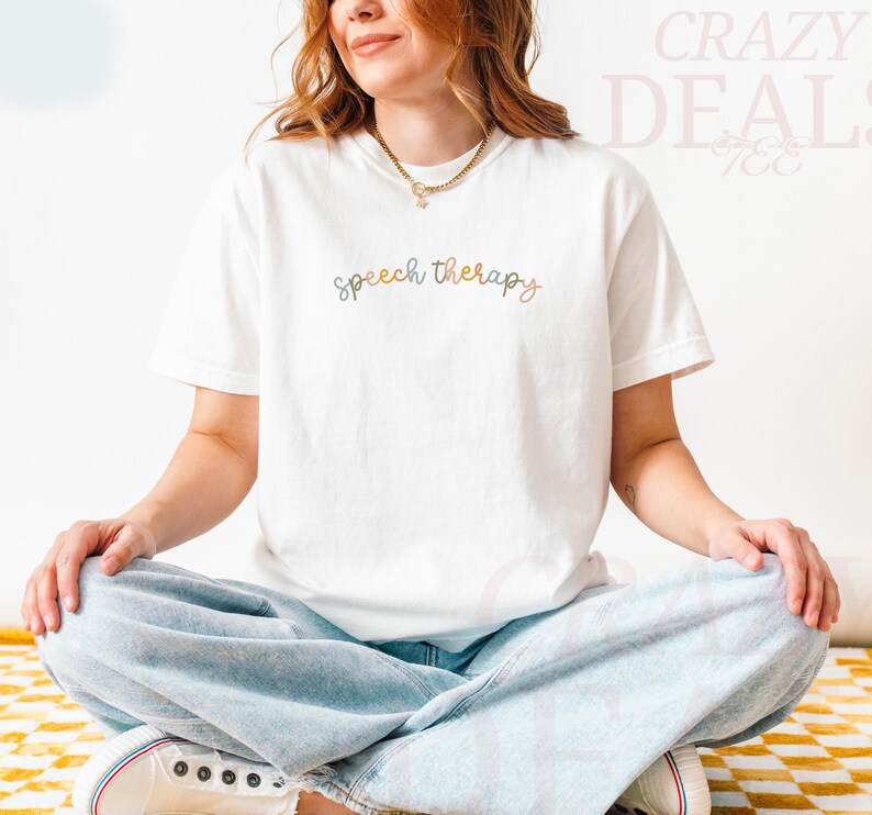 May include: White t-shirt with the words "speech therapy" in a cursive, multi-colored font. The shirt is worn with light blue jeans and white sneakers. The person is sitting cross-legged on a yellow and white checkered surface.