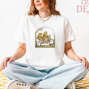 May include: White t-shirt featuring a whimsical illustration of two frogs riding a tandem bicycle. The artwork is framed by an archway with foliage. The words "CRAZY DEALS" are printed in pink on the upper right.