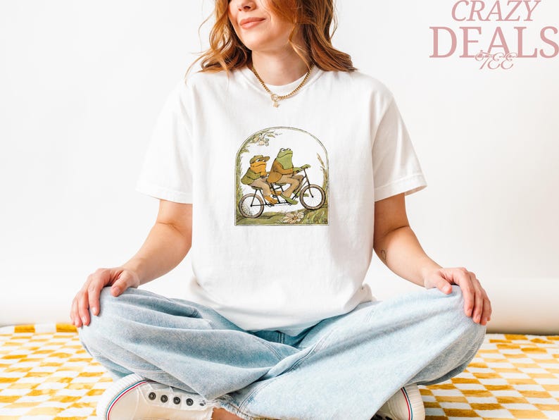 May include: White t-shirt featuring a whimsical illustration of two frogs riding a tandem bicycle. The artwork is framed by an archway with foliage. The words "CRAZY DEALS" are printed in pink on the upper right.