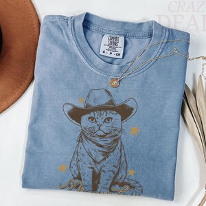 May include: A light blue t-shirt featuring a graphic of a cat wearing a cowboy hat and bandana. The cat is surrounded by gold stars and a rope design. The shirt has a Comfort Colors tag.